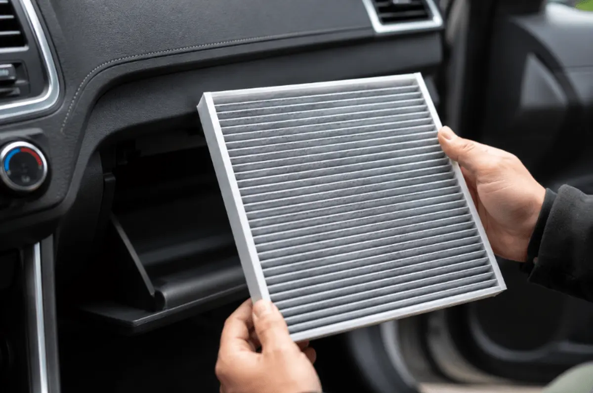 Hands holding a pleated cabin air filter next to a car’s open glove box.